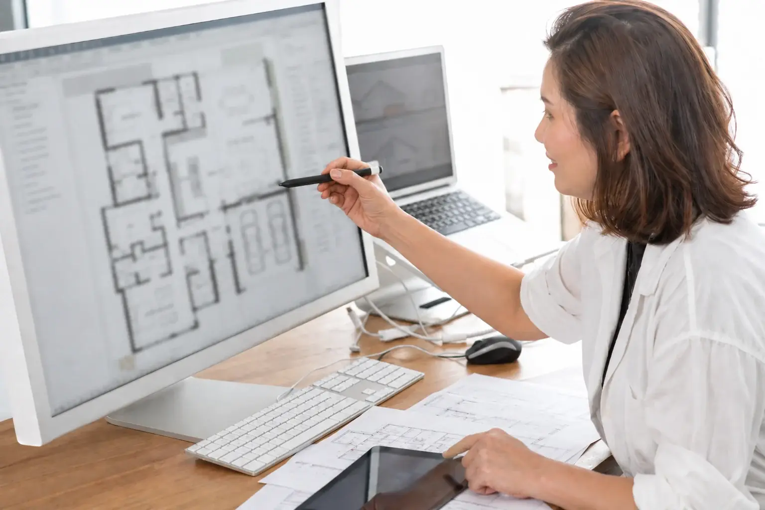 An architect at a desk pointing a pen at a house floor plan on a large screen, with printed plans beside the keyboard