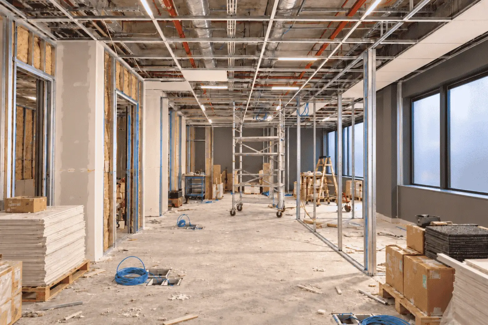 An office fit-out under construction: steel stud walls with insulation, an open ceiling grid with ducts and sprinkler pipes, and data cables coiled at the floor boxes