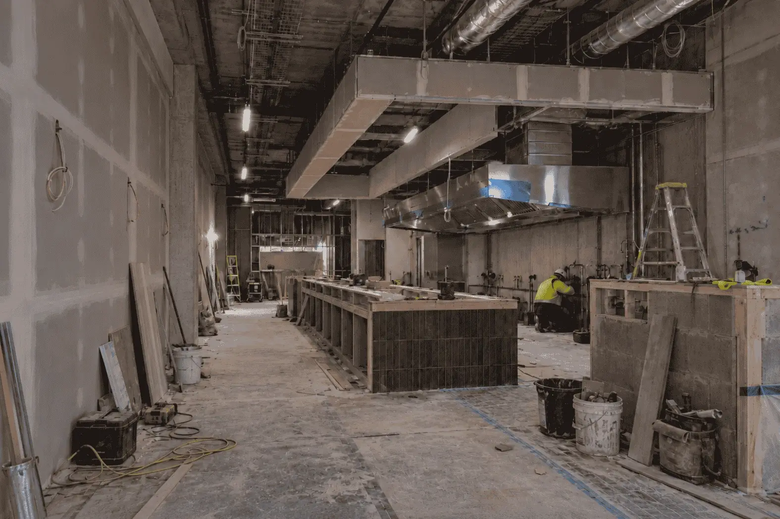 A restaurant fit-out under construction: a stainless-steel kitchen exhaust canopy in place, a tiled counter frame half built and a plumber at the kitchen pipework