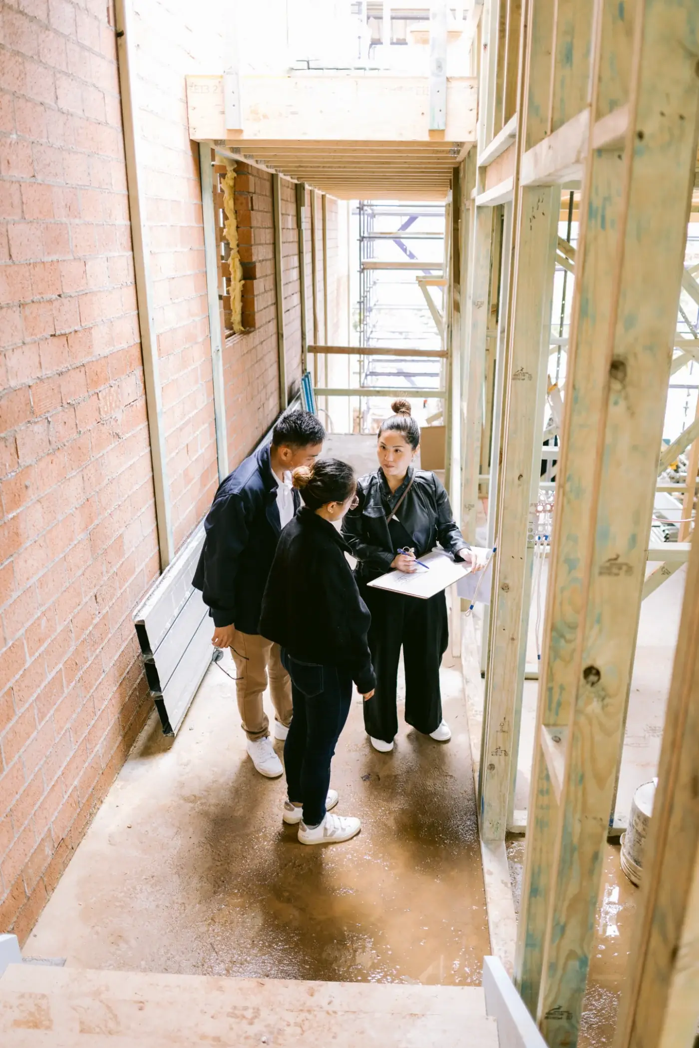 An OIC architect going over plans with two clients between timber framing and a brick wall on a building site